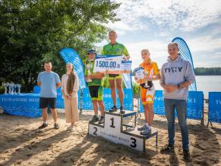 Sześć osób uśmiecha się do zdjęcia na tle jeziora — od lewej: mężczyzna, kobieta, trzy osoby na podium z nagrodami i mężczyzna.