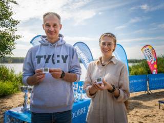 Mężczyzna i kobieta uśmiechają się do zdjęcia i trzymają odznakę "Tyska Pętla Rowerowa". W tle widać puchary, które leżą na stoliku i jezioro.