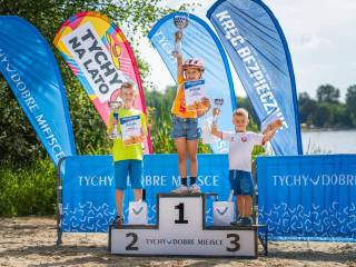 Troje dzieci stoi na podium z pucharami i nagrodami, w tle baner z napisem "Tychy dobre miejsce" i jezioro.
