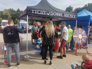 Dorośli z dziećmi stoją przy namiocie z napisem "Tychy dobre miejsce". W tle widać parking, karetkę pogotowia, radiowóz policyjny i las.