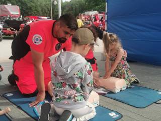 W otwartym namiocie dwóch ratowników medycznych w odblaskowych ubraniach tłumaczy chłopcu i dziewczynce, jak udzielać pierwszej pomocy.