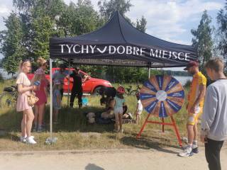 W otwartym namiocie dwóch ratowników medycznych w odblaskowych ubraniach tłumaczy chłopcu i dziewczynce, jak udzielać pierwszej pomocy. Obok stoją ludzie, a w tle widać czerwony samochód, drzewa i jezioro.