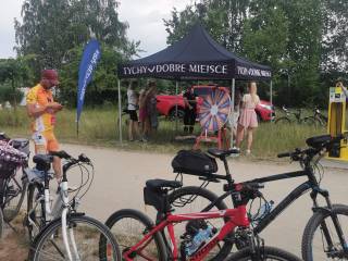 Przy czarnym namiocie plenerowym z napisem "Tychy dobre miejsce" stoją ludzie. Z przodu znajdują się rowery i kolarz, który patrzy w telefon. W tle widać czerwony samochód i jezioro.