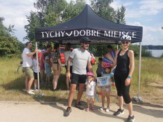 Mężczyzna, kobieta i dwie dziewczynki stoją w kaskach i uśmiechają się do zdjęcia. Za nimi znajdują się: namiot plenerowy, ludzie, czerwony samochód i jezioro.