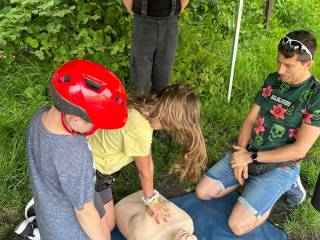 Chłopiec i dziewczynka uczą się resuscytacji na manekinie pod opieką dwóch dorosłych mężczyzn. Wszystko odbywa się na świeżym powietrzu