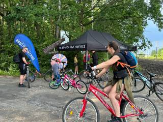 Kobieta jedzie na czerwonym rowerze. W tle stoją dwaj mężczyźni oraz troje dzieci z rowerami. Za nimi znajduje się czarny namiot plenerowy i las.