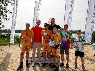 Czterech mężczyzn, kobieta i dwóch chłopców w strojach kolarskich stoją na tle podium i jeziora.