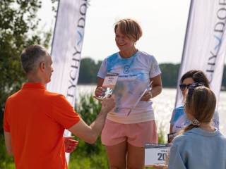 Kobieta odbiera nagrodę od mężczyzny w pomarańczowej bluzce. Obok stoją dwie kobiety. W tle widać jezioro i drzewa.