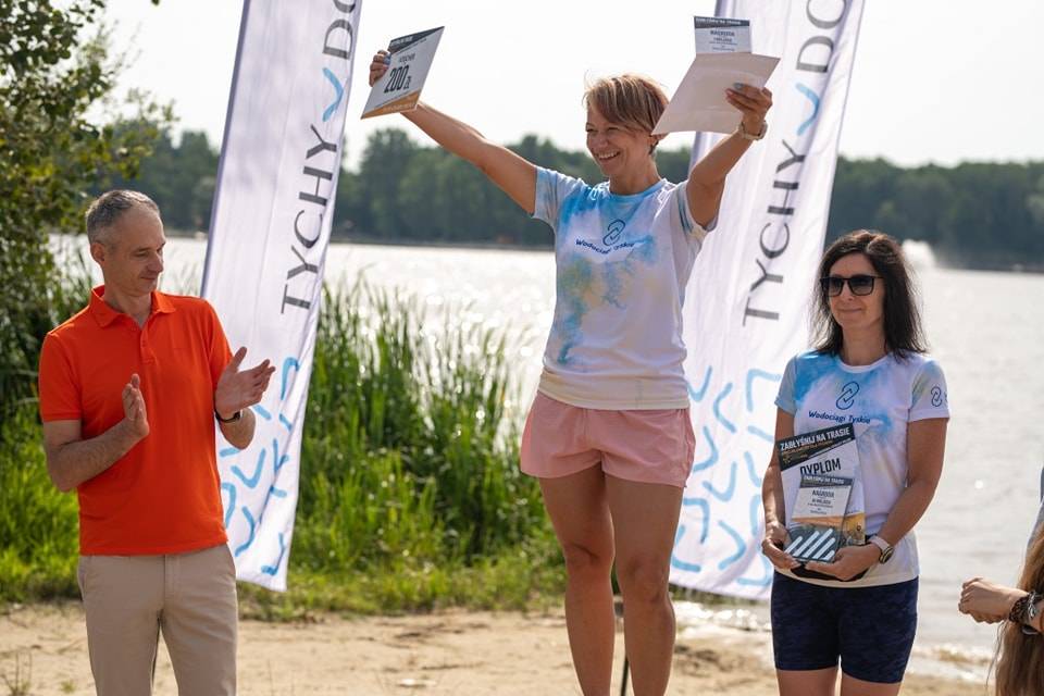 Dwie kobiety z nagrodami stoją obok mężczyzny bijącego brawo. W tle widać jezioro.