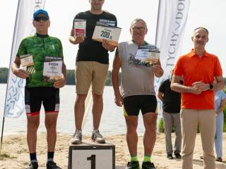 Trzech mężczyzn stoi na podium z nagrodami, obok nich stoi mężczyzna. W tle widać jezioro.