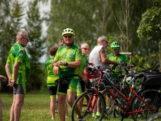 Grupa ludzi w zielonych strojach stoi przy rowerach. W tle znajduje się las.