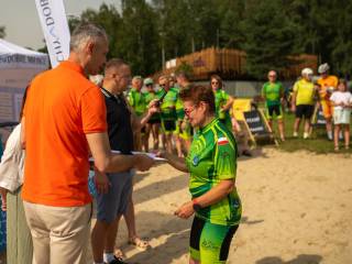 Kobieta w zielonym stroju sportowym odbiera od mężczyzny kartkę, obok nich i w tle stoją ludzie, widać także biały namiot z napisem "Tychy dobre miejsce".  