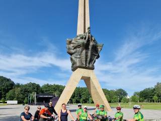 Grupa ludzi z rowerami pozuje do zdjęcia. W tle Pomnik Walki i Pracy, scena i drzewa.