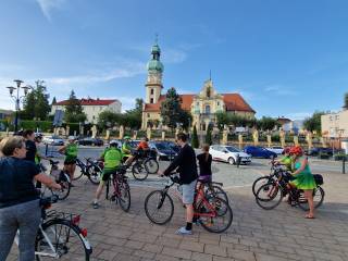 Na brukowanej drodze znajduje się grupa ludzi na rowerach. Za nimi widać kościół.