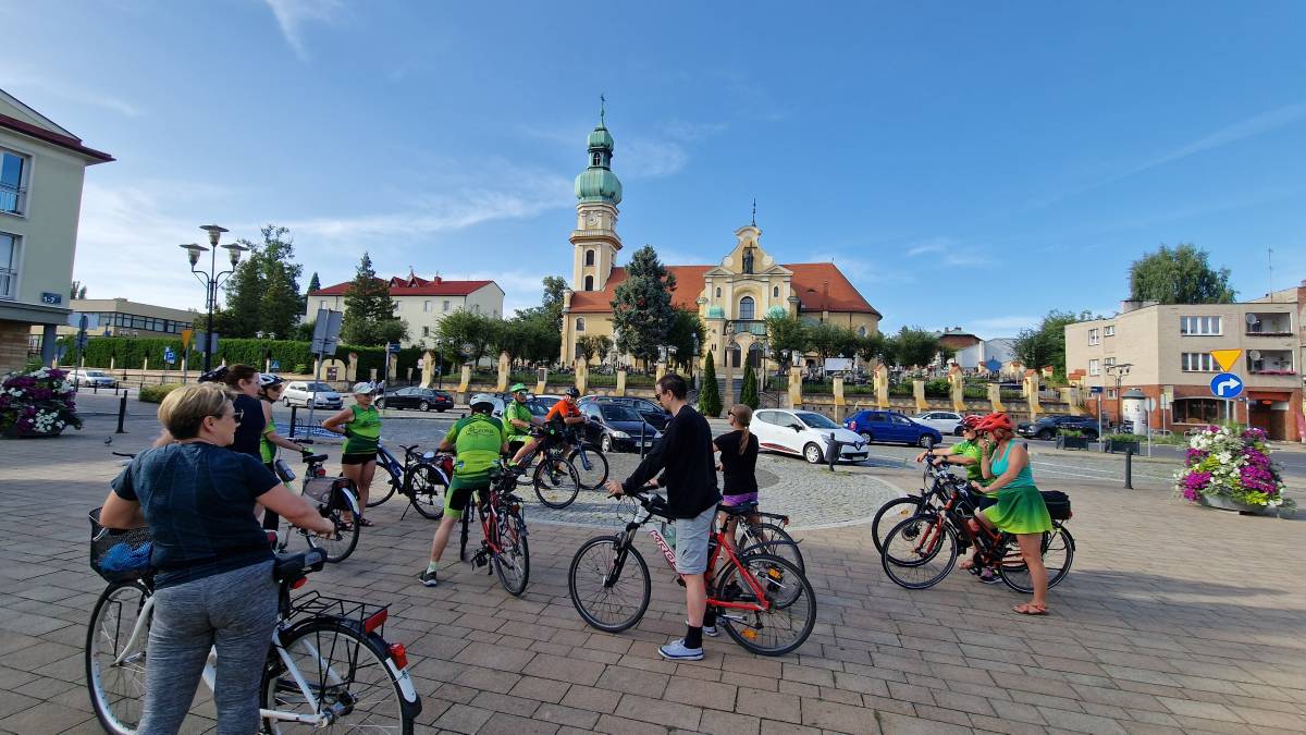 Na brukowanej drodze znajduje się grupa ludzi na rowerach. Za nimi widać kościół.