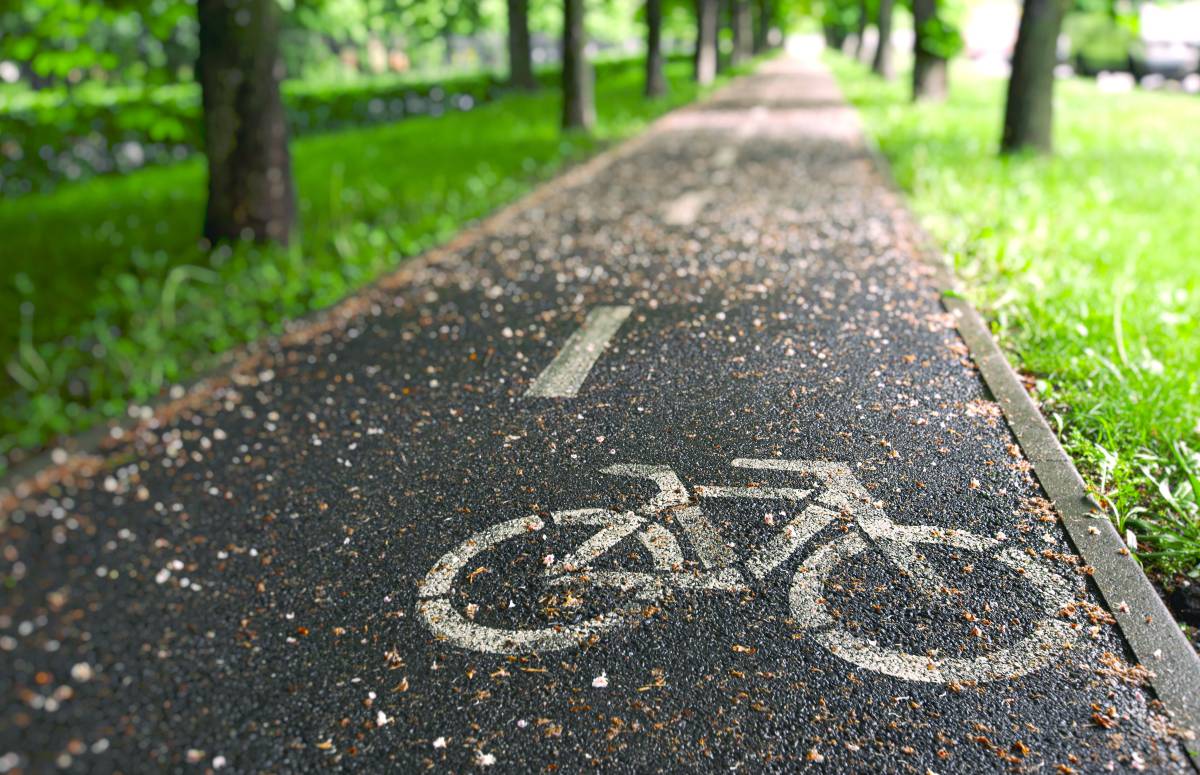 Droga rowerowa oznaczona białym symbolem roweru. Na nawierzchni leżą liście, po bokach ścieżki jest zielona trwa i drzewa.