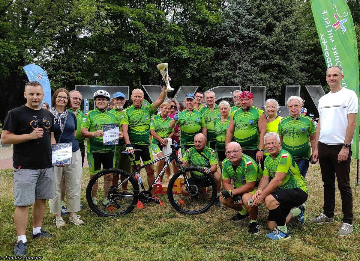 Duża grupa ludzi w sportowych strojach uśmiecha się do zdjęcia, w tle napis "Tychy" i zielone drzewa.