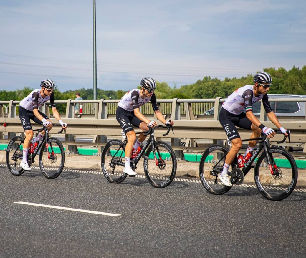 Trzech kolarzy w sportowych strojach jedzie po asfaltowej drodze podczas wyścigu.