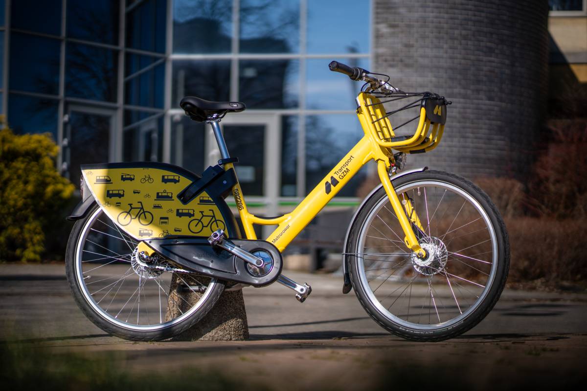 Żółty rower z napisem "Transport GZM" na tle przeszklonego budynku.