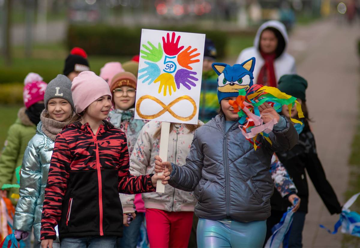 Dwie dziewczynki trzymają baner z namalowanymi kolorowymi dłoniami, za nimi idzie grupa dzieci.