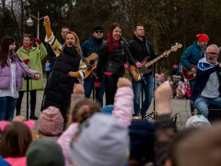 Trzy kobiety i czterech mężczyzn śpiewają i grają na instrumentach na placu w parku, w tle tańczą ludzie.
