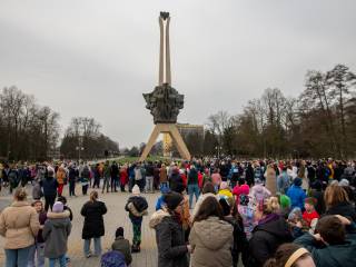 Duża grupa ludzi stoi na placu przy pomniku w parku, w tle budynek i drzewa.