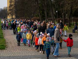 Grupa dzieci idzie z nauczycielami po chodniku w parku, w tle widać drzewa i budynek