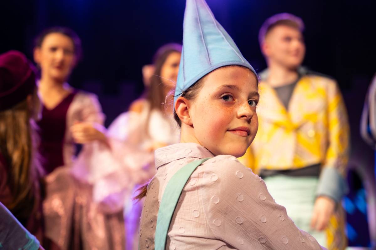 Dziewczynka w niebieskiej czapce i różowej koszuli uśmiecha się do zdjęcia, w tle widać ludzi.