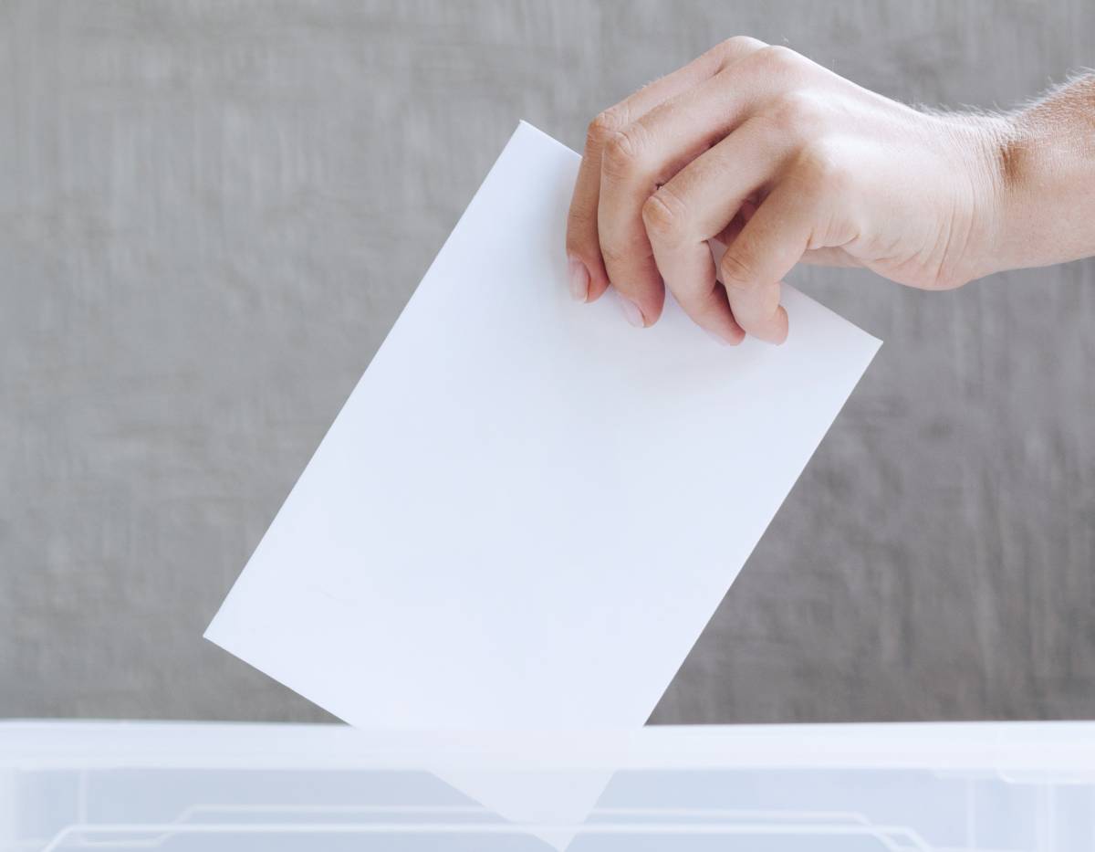 Dłoń wrzuca kartę do urny wyborczej na tle szarej ściany