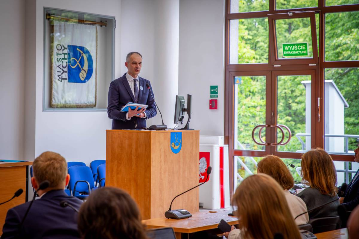 Mężczyzna w garniturze wygłasza przemówienie,w tle widoczny herb miasta.