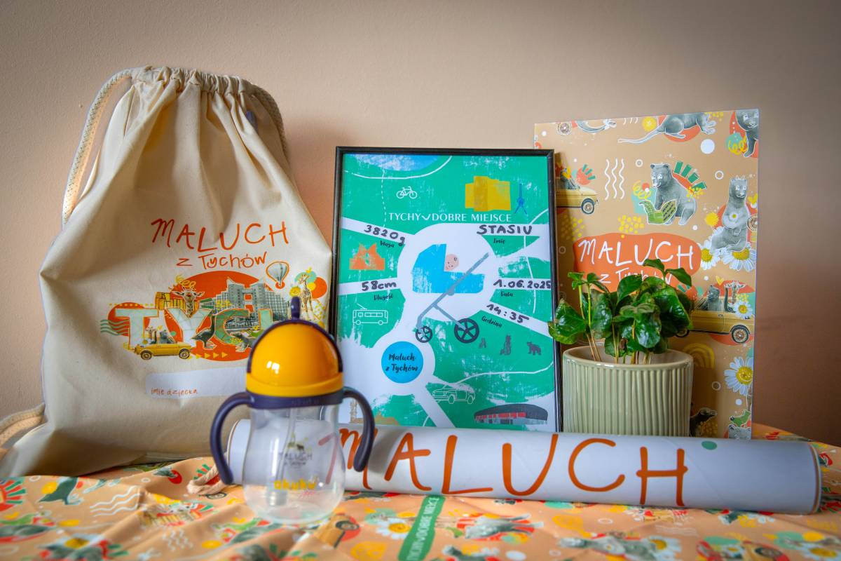 Na tle kremowej ściany leżą na chuście z symbolami Tychów: worek z napisem ''Maluch z Tychów'', butelka dla noworodka, kwiatek w doniczce, metryczka, teczka oraz miarka w tubie z napisem "Maluch".