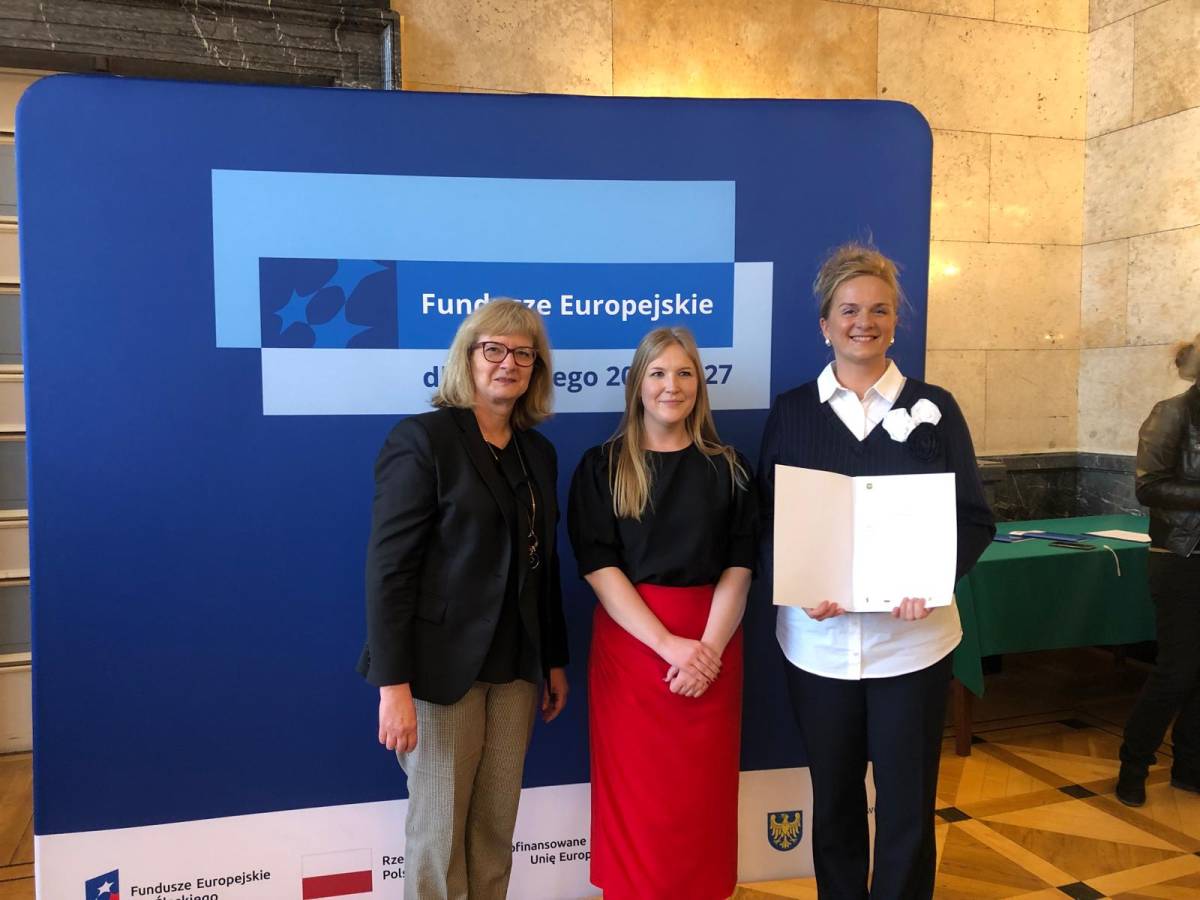 Trzy kobiety stoją na tle niebieskiej ścianki z napisem "Fundusze Europejskie" i uśmiechają się do zdjęcia.