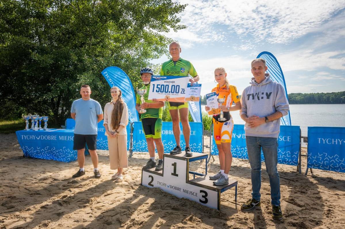 Sześć osób uśmiecha się do zdjęcia na tle jeziora — od lewej: mężczyzna, kobieta, trzy osoby na podium z nagrodami i mężczyzna.