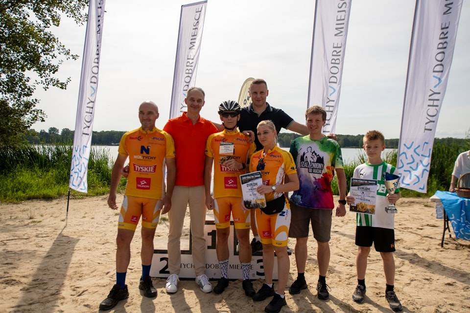 Czterech mężczyzn, kobieta i dwóch chłopców w strojach kolarskich stoją na tle podium i jeziora.