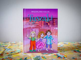 Różówa książka "Tyszaki – Magdalena Kulus" z grafiką trzech dziewczynek (tyszaków), leży na pomarańczowej chuście z symbolami miasta. W tle kremowa ściana.