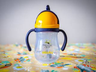 Butelka niekapek z granatowym uchwytem i żółtą zakrętką, przezroczysta z napisem "Maluch z Tychów, akuku", leży na pomarańczowej chuście ozdobionej symbolami miasta, na tle kremowej ściany.