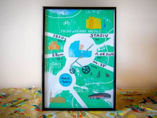 Metryczka w zielonym kolorze, oprawiona w czarną ramkę z grafiką niemowlęcia w niebieskim wózku, leży na pomarańczowej chuście z miejskimi symbolami W tle kremowa ściana.