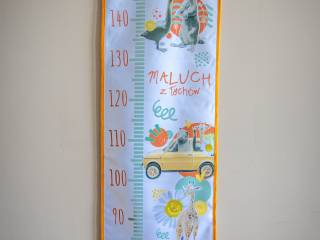 Miarka biało-pomarańczowa z kolorowymi symbolami miasta i napisem "Maluch z Tychów" na tle kremowej ściany.