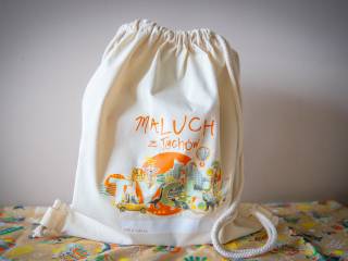 Lniany beżowy worek z pomarańczowym napisem "Maluch z Tychów", grafiką symboli miasta oraz napisem "Tychy". Leży na pomarańczowej chuście z symbolami miasta. W tle kremowa ściana.
