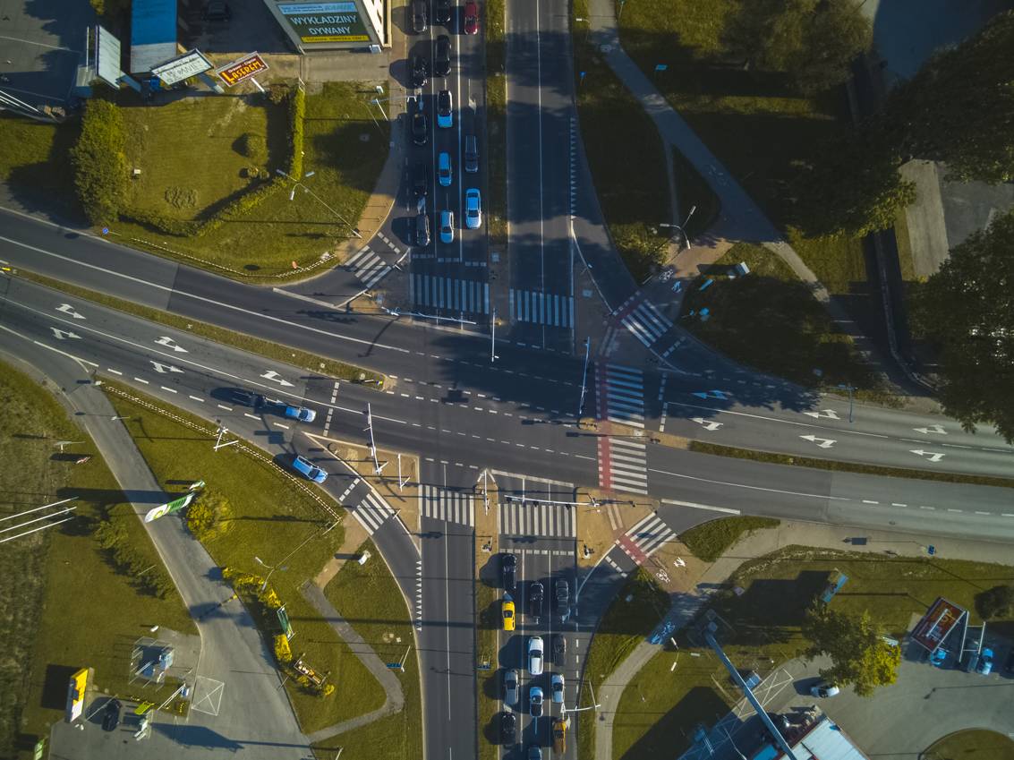 Widok z drona na skrzyżowanie — widoczne są auta, ulice i trawniki.