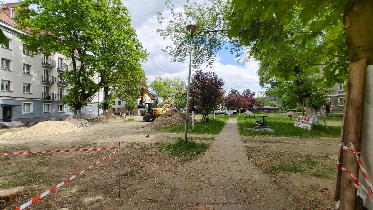 Plac i chodnik w trakcie prac remontowych, w tle bloki mieszkalne oraz drzewa.