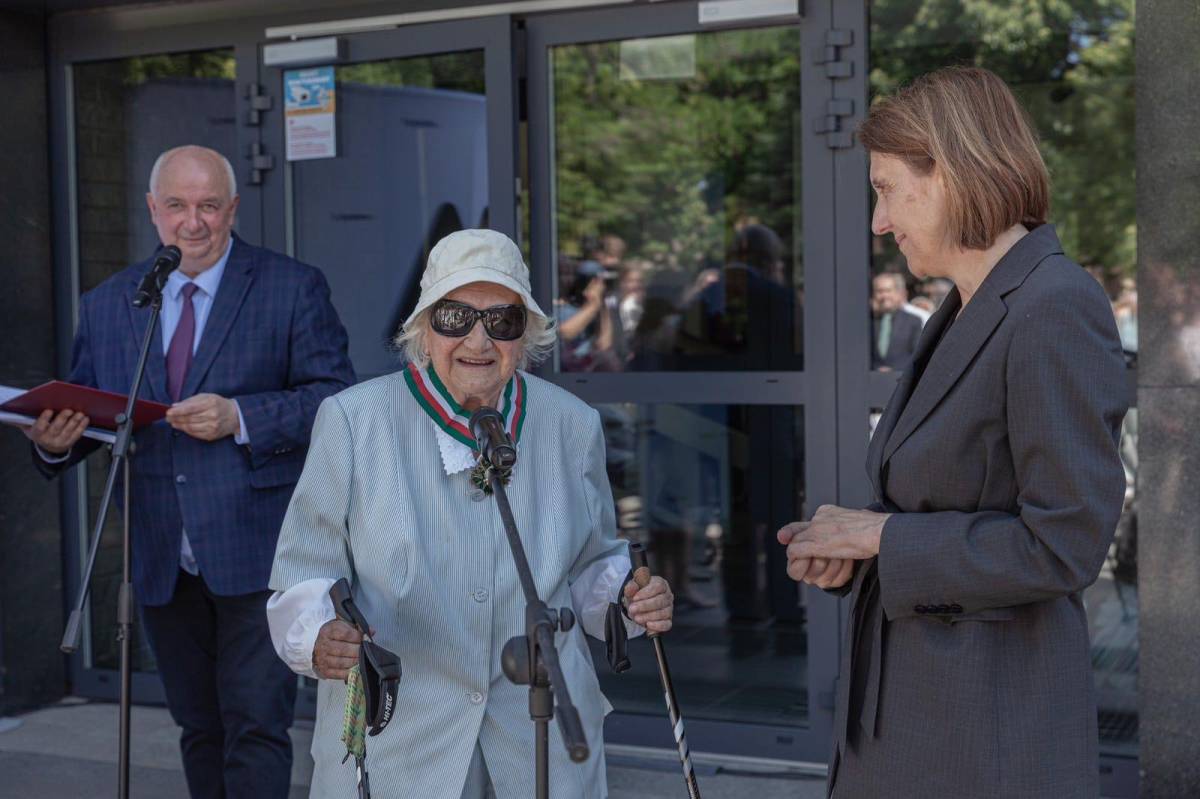 Dwie kobiety stoją obok siebie przed mikrofonem – młodsza w marynarce i starsza, elegancko ubrana z medalem na szyi, uśmiechają się. W tle widać mężczyznę przemawiającego do mikrofonu oraz fragment przeszklonego budynku, w którym odbijają się sylwetki ludzi.