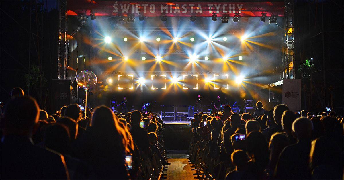 Pod sceną stoi duża grupa osób, miejsce rozświetlają reflektory.