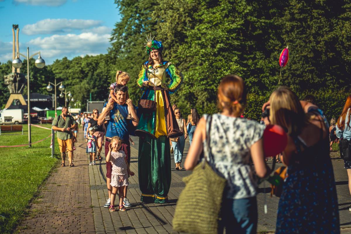 Kolorowo ubrana kobieta stoi na szczudłach obok mężczyzny i dwójki dzieci. Wszyscy uśmiechają się do zdjęcia, które robią dwie kobiety stojące przed nimi. W tle widać pomnik, ludzi oraz drzewa.