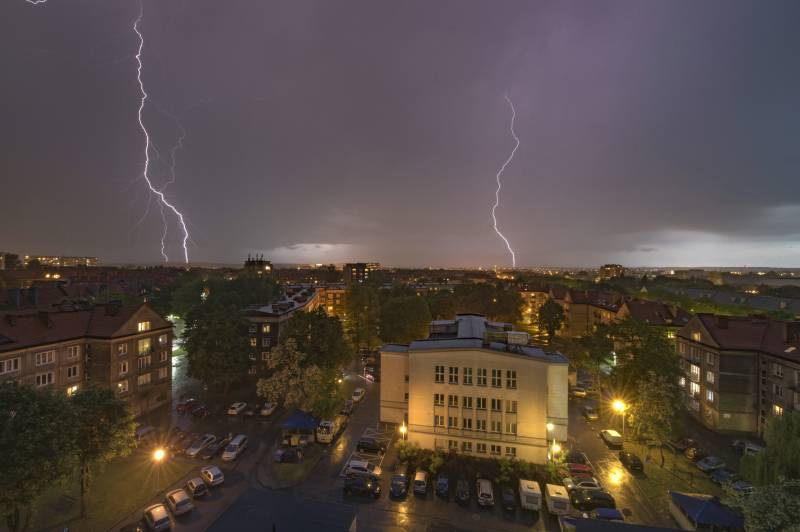 Widok z drona na miasto podczas burzy: widać bloki mieszkalne, ulicę i dwie błyskawice rozświetlające niebo.