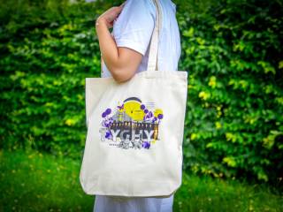 Kobieta stoi w białej koszulce, na ramieniu trzyma lnianą kremową torbę z napisem "Tychy", w tle widać zielone krzewy.