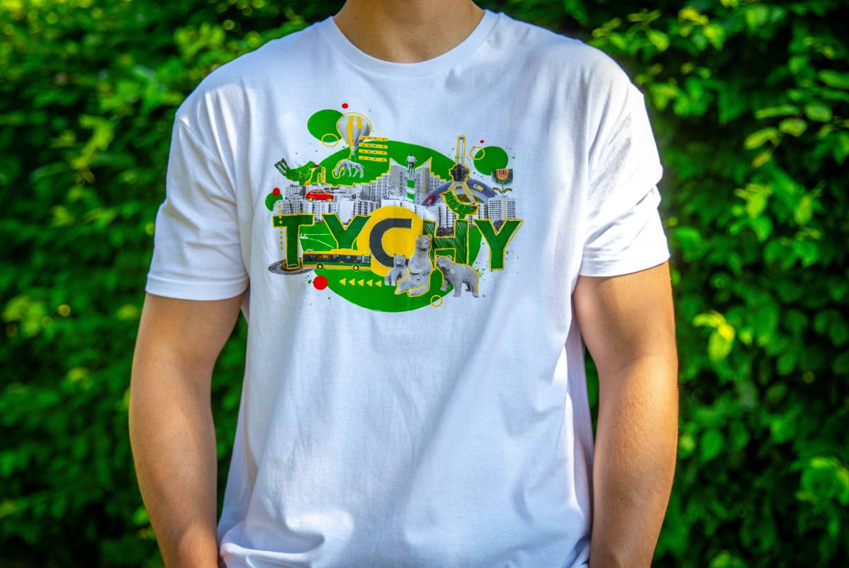 Mężczyzna stoi w białej koszulce z zielonym nadrukiem, symbolami miasta i napisem "Tychy", w tle widać zielone krzewy.