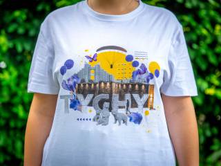 Kobieta stoi w białej koszulce z zielonym nadrukiem, symbolami miasta i napisem "Tychy", w tle widać zielone krzewy.