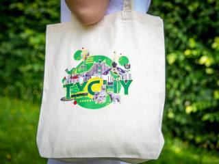 Kobieta stoi w białej koszulce, na ramieniu trzyma lnianą kremową torbę z napisem "Tychy", w tle widać zielone krzewy.