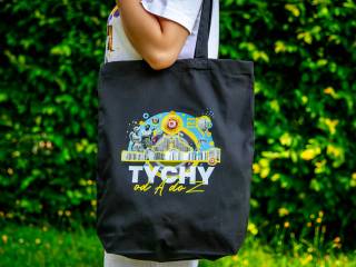 Kobieta stoi w białej koszulce, na ramieniu trzyma lnianą czarną torbę z napisem "Tychy", w tle widać zielone krzewy.